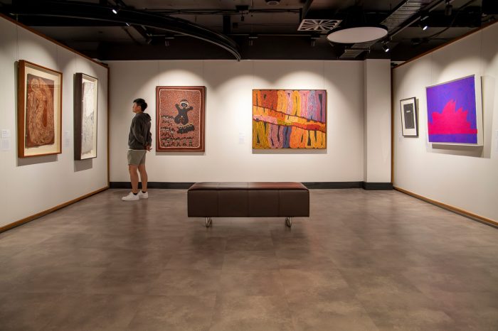 A wide view of an art gallery room with six paintings hanging on the white walls. On the left of the image a man stands admiring an artwork. 