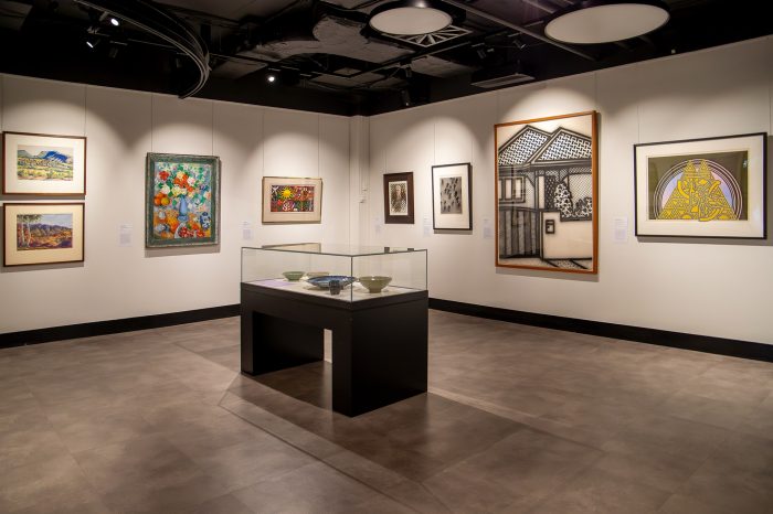 A wide view of an art gallery room with eight paintings hanging on the white walls. In the middle of the room is a glass display table containing five ceramic bowls. 