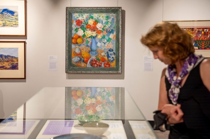 A woman stands in front of a glass display table, viewing a selection of ceramic bowls. In the background, and in focus, is a colourful painting depicting flowers and fruit on a table. 