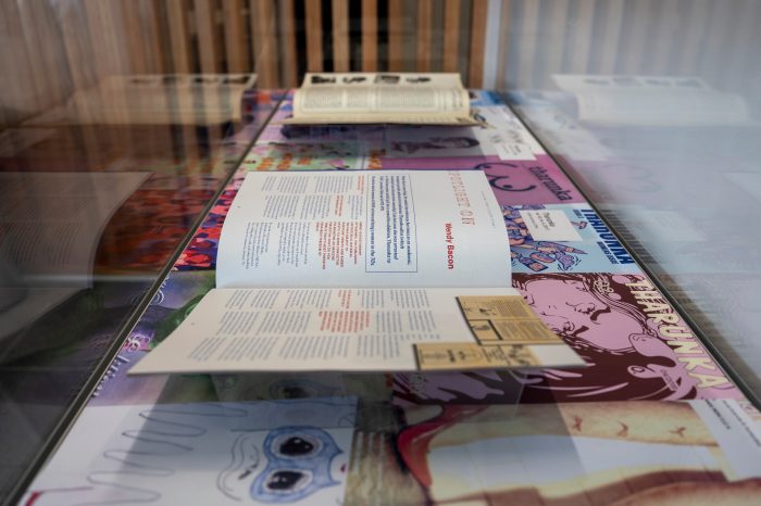 A side-angled view of a glass display cabinet, containing two opened magazines. Text on the closest magazine reads "Spotlight on Wendy Bacon". 