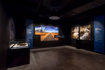 A dark room with three display walls. The middle wall displays imagery of an orange desert against a blue sky. The right wall displays two large photos of an underground cave. 