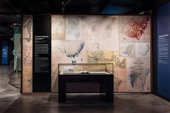 A display case sits in front of a wall covered in a collage of archival maps of Australia