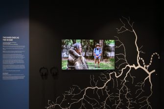 A black wall with a TV screen showing woman wearing a possum pelt cape, addressing people in a park. On the black wall is a white schematic of a river system. 