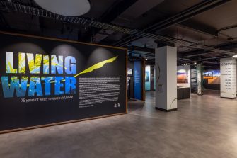 View of a large exhibition space. To the left, closest to the camera is a black wall with bright blue text reading "Living Water". In the distance are hints of colourful displays on the walls and in display cases. 