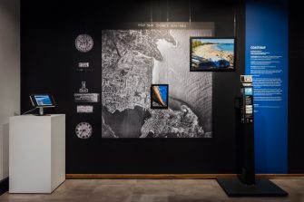 A wall of a exhibition with a large black and white aerial photo of a coastline. On top of the aerial photo sits two colour images in frames, also depicting the coastline from different angles. 