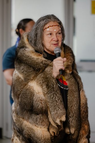 A women wearing a fur cape holds a microphone, addressing the audience