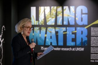 A lady with white hair holds a microphone in one hand, and a piece of paper in another hand. In the background is a wall that says "Living Water".