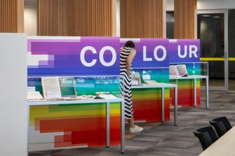 An image of an exhibit with three display cases, and a large panel displaying the word "COLOUR" in colourfully designed blocks. A woman is looking at a display case filled with books and other items related to colour..