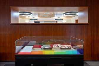 The image shows a glass display case containing a collection of colour swatches. The display case is situated in a room with wood-panelled walls and circular lights overhead.