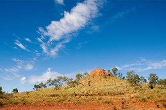 Revealling Riversleigh - hero 01 A vast, semi-arid landscape revealing orange dirt, green shrubbery, and a small hill peaking with a cluster of large rocks.